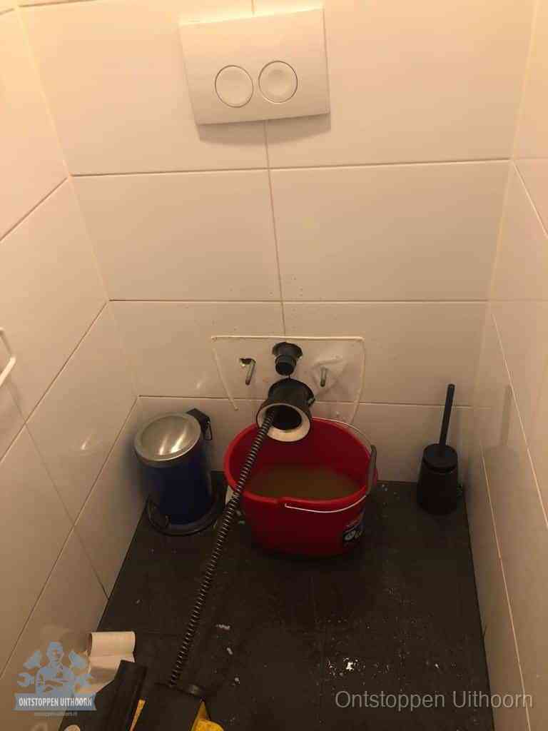 Een ontstopte wand-wc met verwijderde toiletpot waar een ontstoppingsveer in de wanduitgang verdwijnt, het achterhalen waar de verstopping zit in Uithoorn