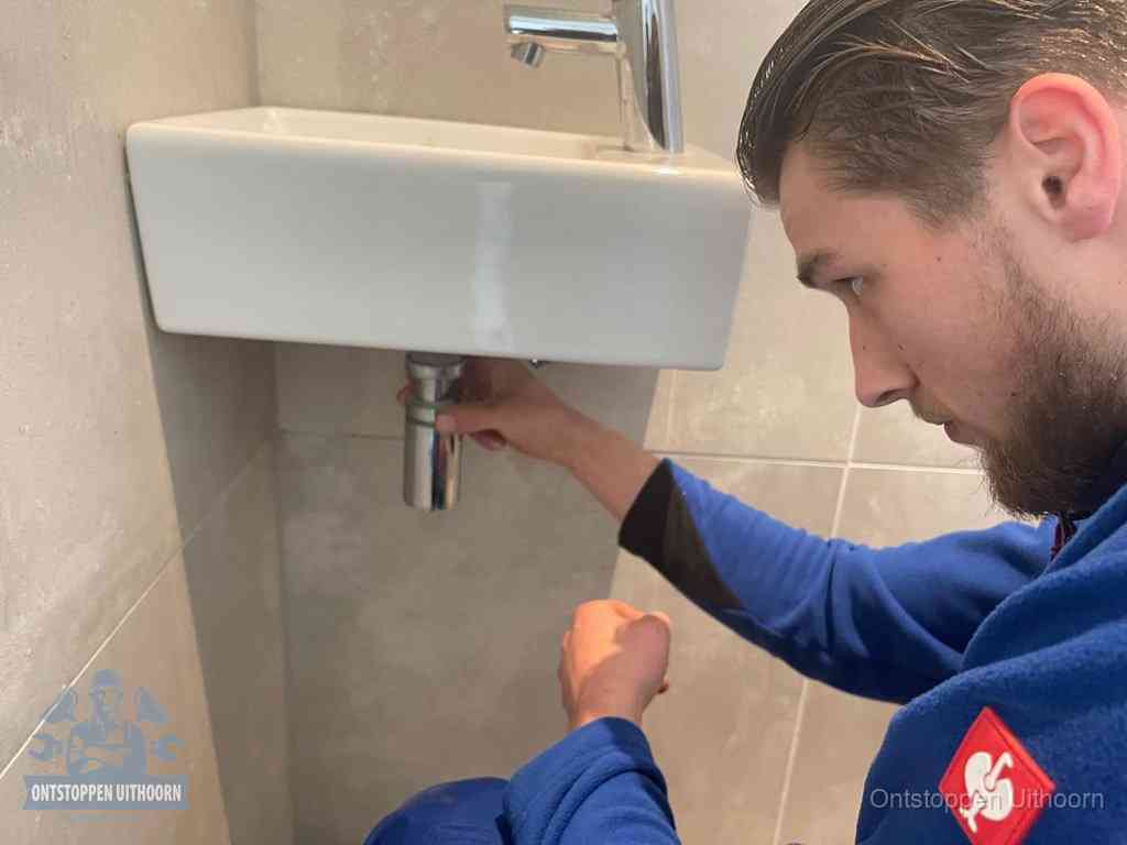 Een monteur draait met de hand de zwanenhalssifon onder een fonteinwasbak in de toiletruimte los om de bron van rioollucht in Uithoorn te vinden