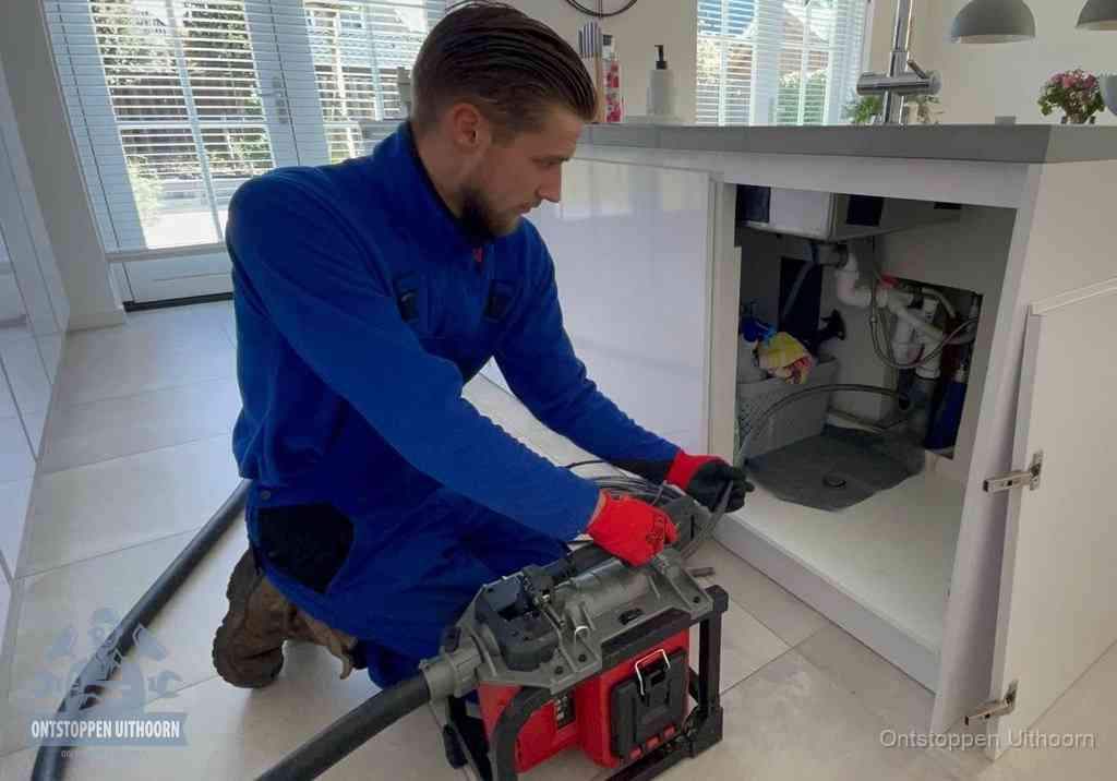 Een monteur voert een professionele elektrische ontstoppingsmachine in de afvoer onder de spoelbak tijdens het gootsteen ontstoppen in Uithoorn
